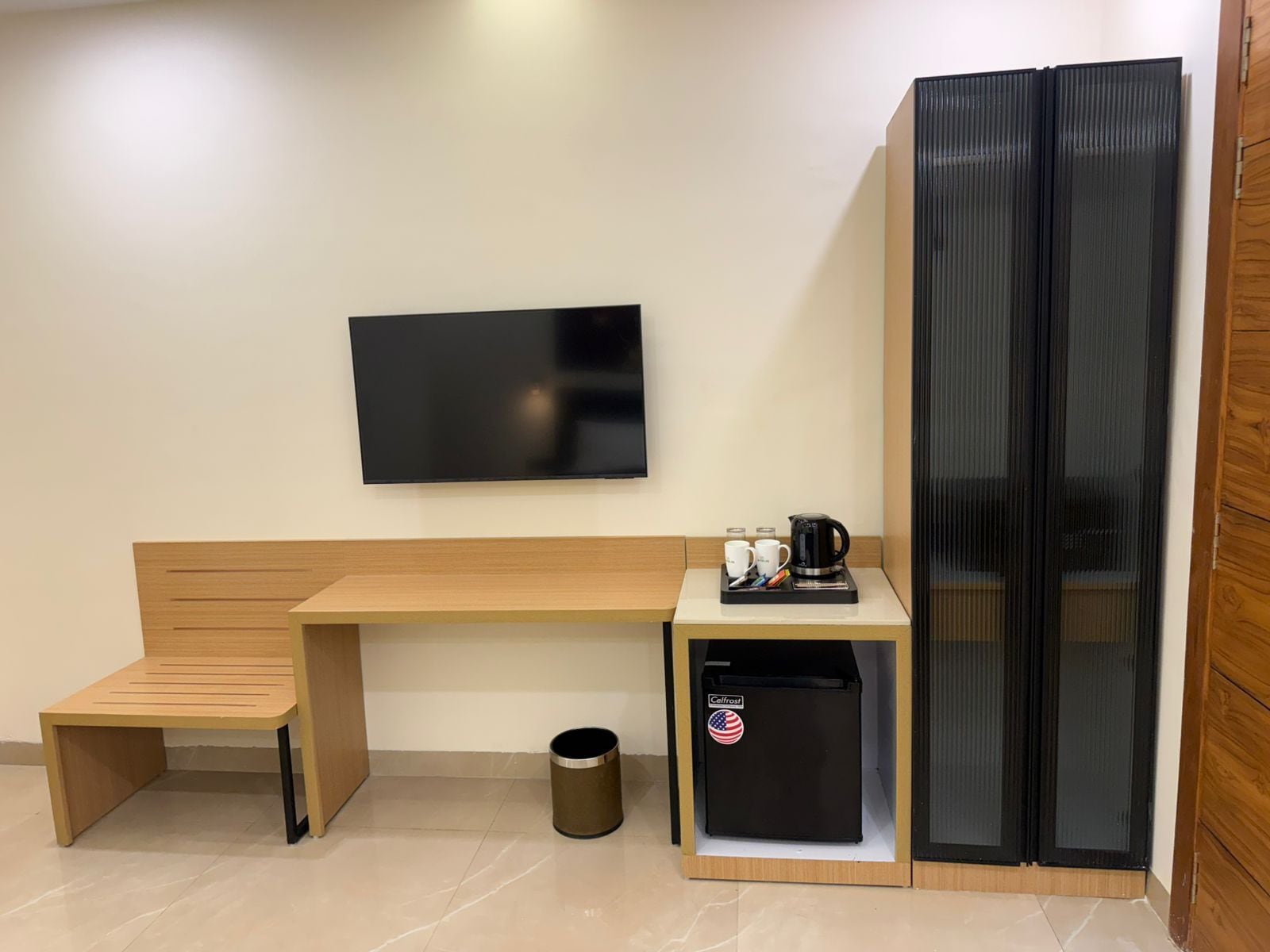 Ein kleines Zimmer mit einer hölzernen Bank, einem schwarzen Kühlschrank, einer Kaffeemaschine und einem Fernseher an der Wand.