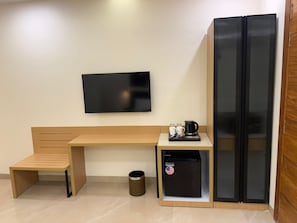 Una habitación pequeña con un banco de madera, un refrigerador negro, una cafetera y un televisor en la pared.