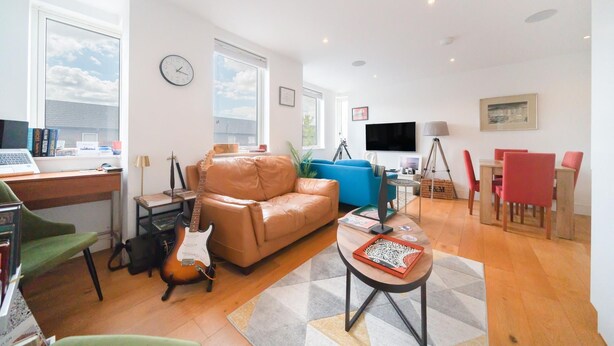 Een moderne woonkamer met een leren bank, een blauwe fauteuil, een eettafel met rode stoelen, een laptop, een gitaar en een flatscreen televisie.