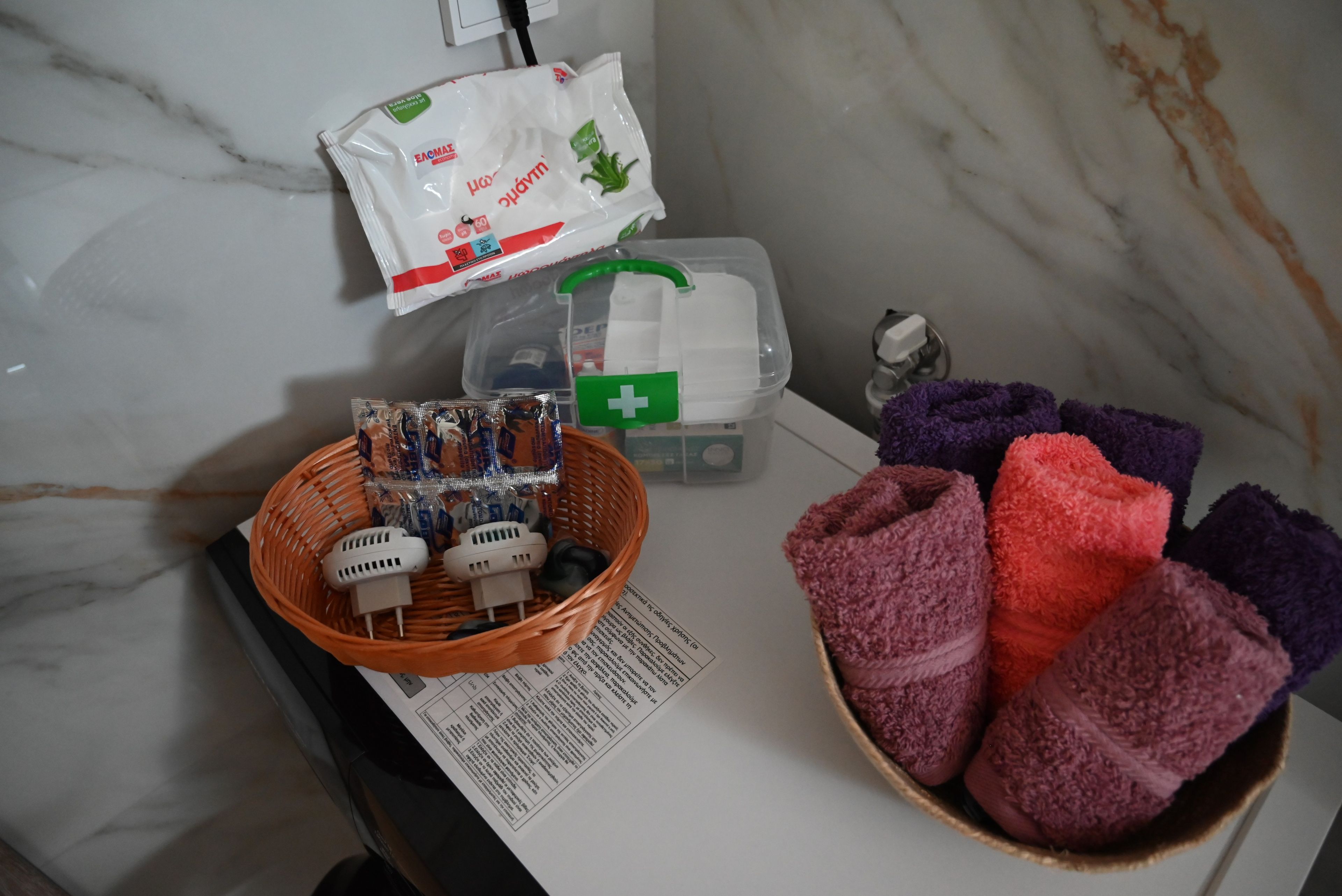 A bathroom with a marble wall, a basket of toiletries, a white bag, a green first aid kit, and a bowl of towels.