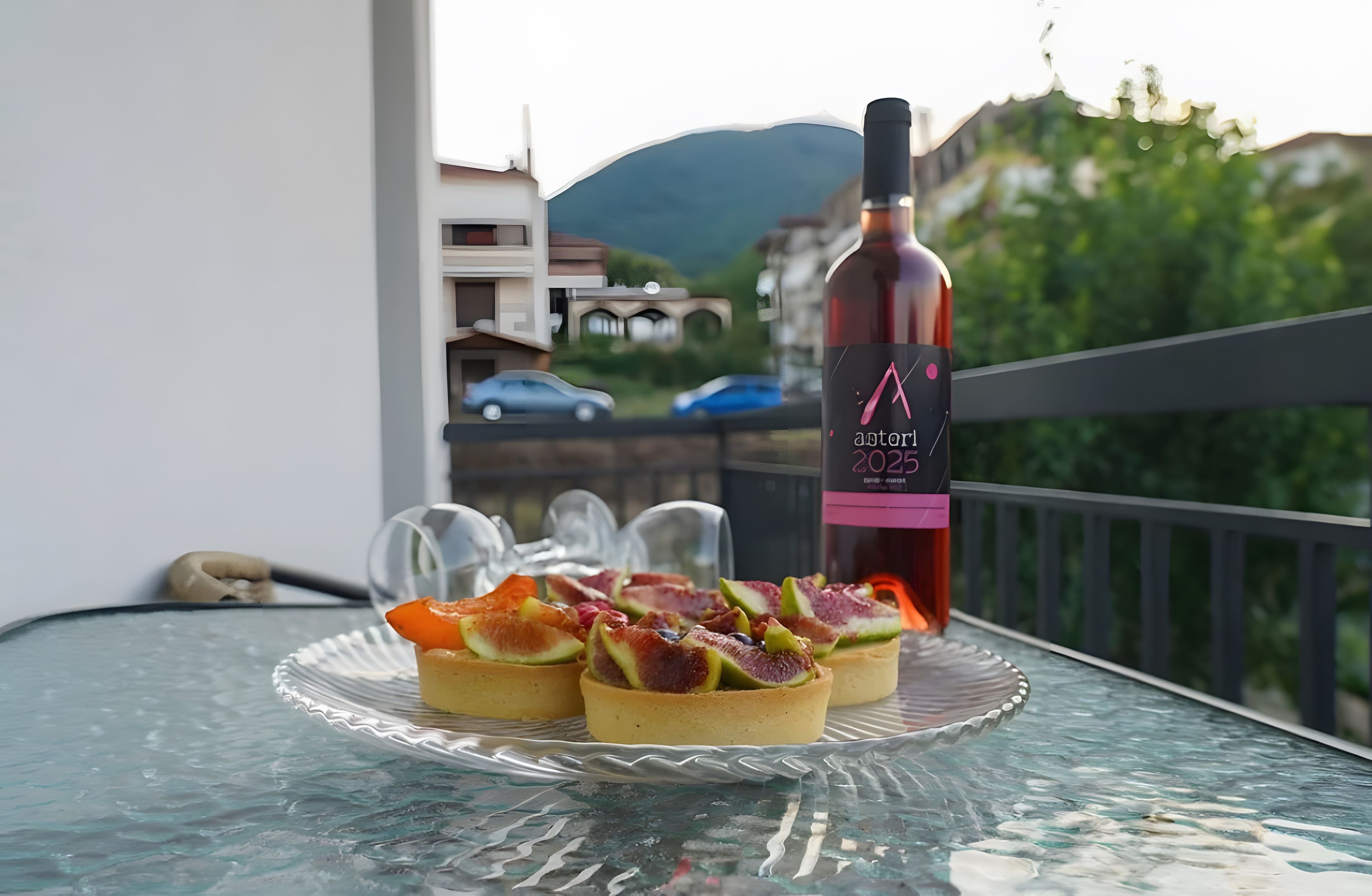 Un tavolino in vetro con una bottiglia di vino e tre dolci su un balcone affacciato su una montagna.