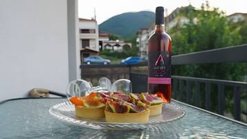 Un tavolino in vetro con una bottiglia di vino e tre dolci su un balcone affacciato su una montagna.