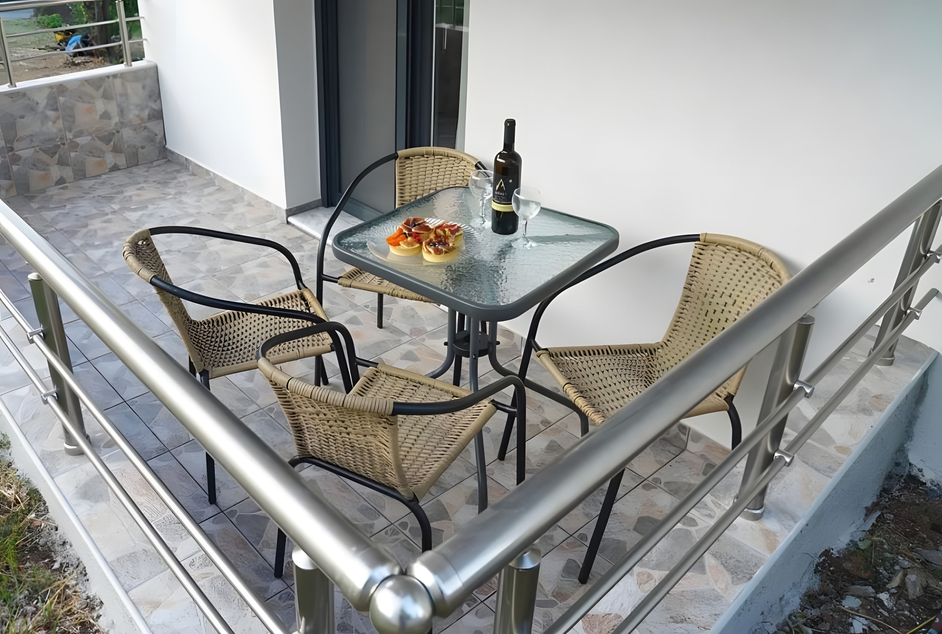 Un balcón con una mesa de vidrio, sillas de mimbre y una botella de vino.
