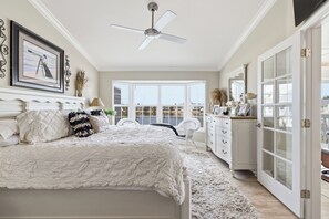 Une chambre avec un grand lit, un ventilateur de plafond, une vue sur l’extérieur grâce à une grande fenêtre et une commode blanche.