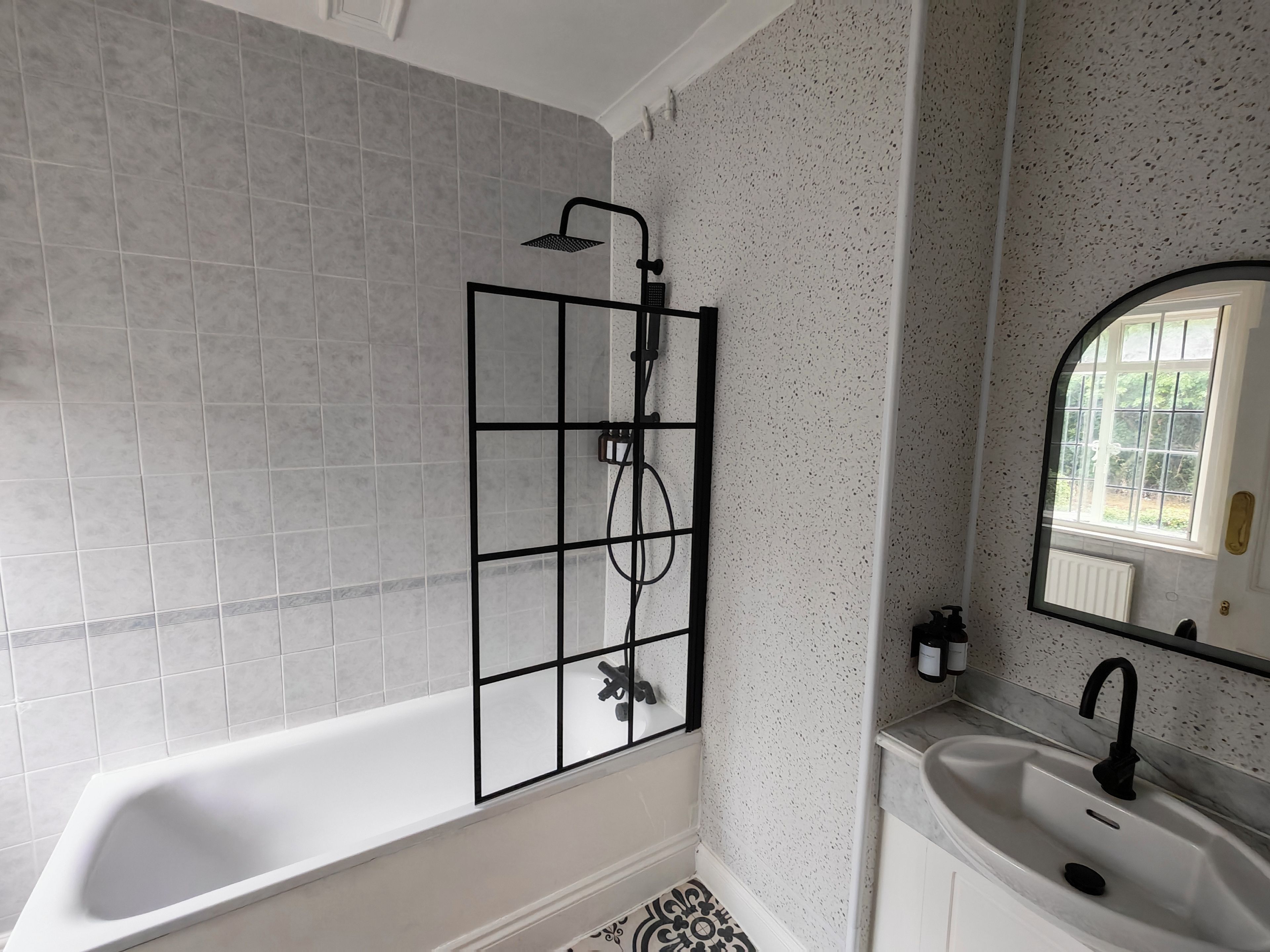 A bathroom with a bathtub, a shower with a black metal frame, a white sink, and a mirror with a window reflection.