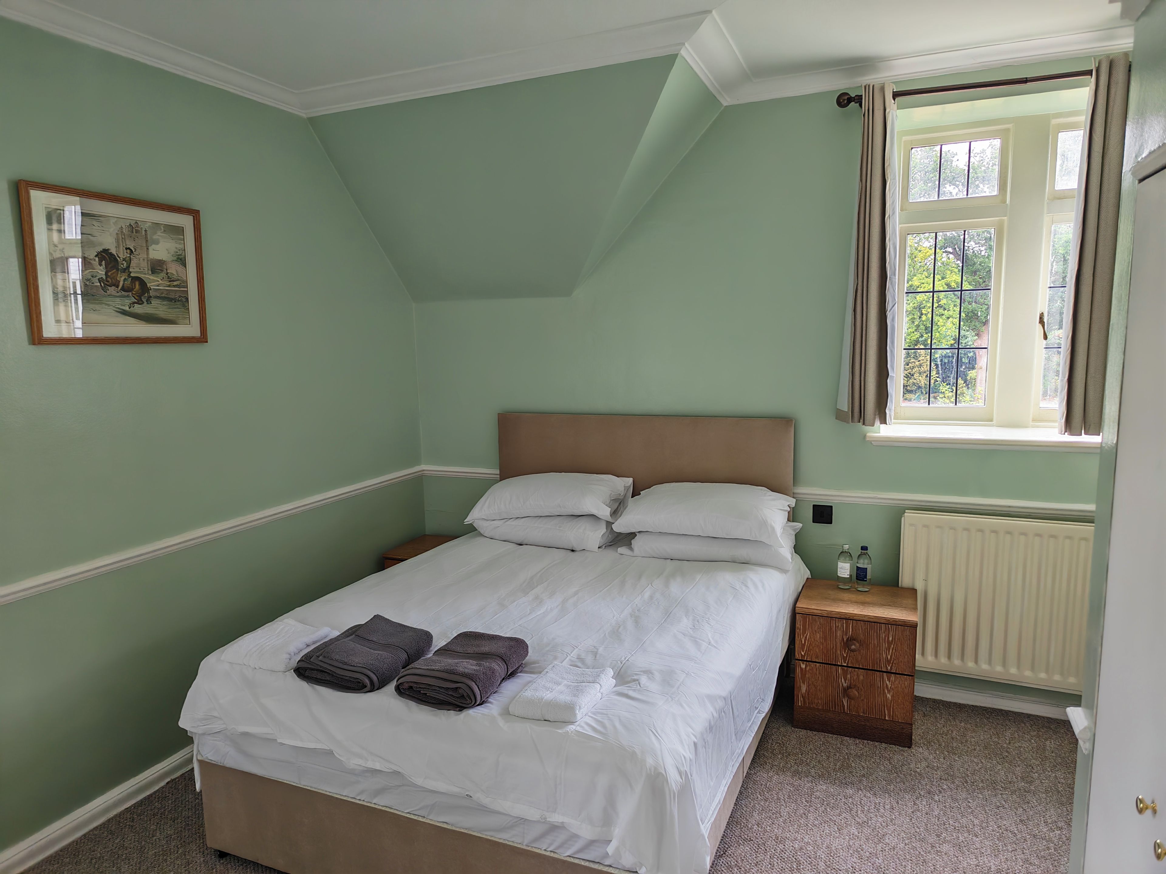 A bedroom with a bed, pillows, a nightstand, a framed picture, a window with curtains, and a radiator.