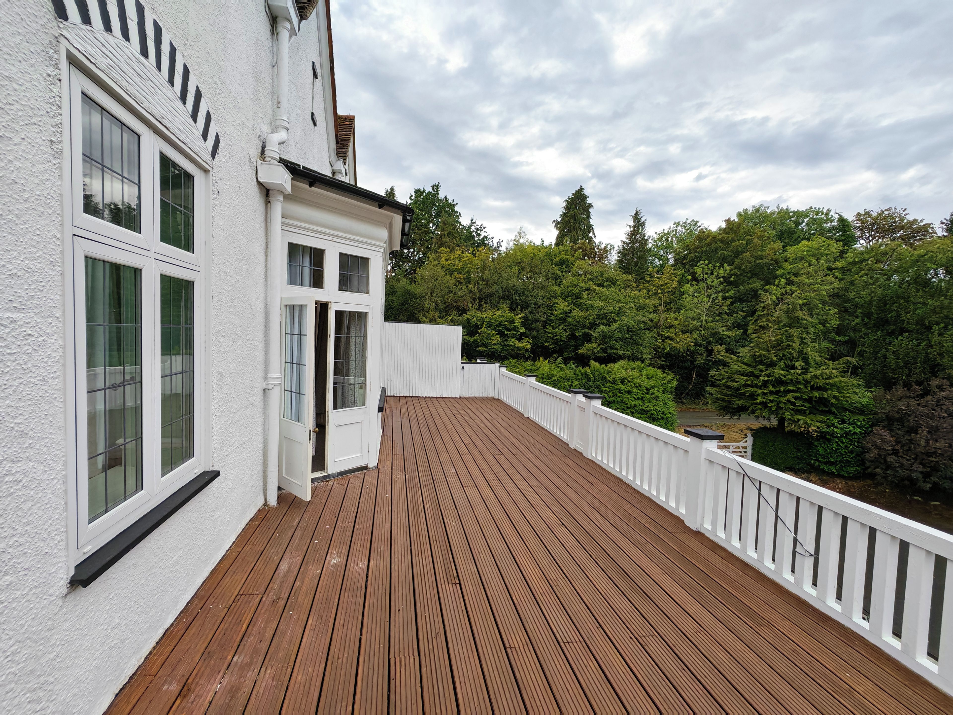Una terraza de madera con barandillas blancas, un edificio con múltiples ventanas y un fondo de árboles.