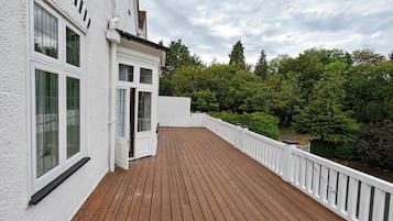 Una terraza de madera con barandillas blancas, un edificio con múltiples ventanas y un fondo de árboles.