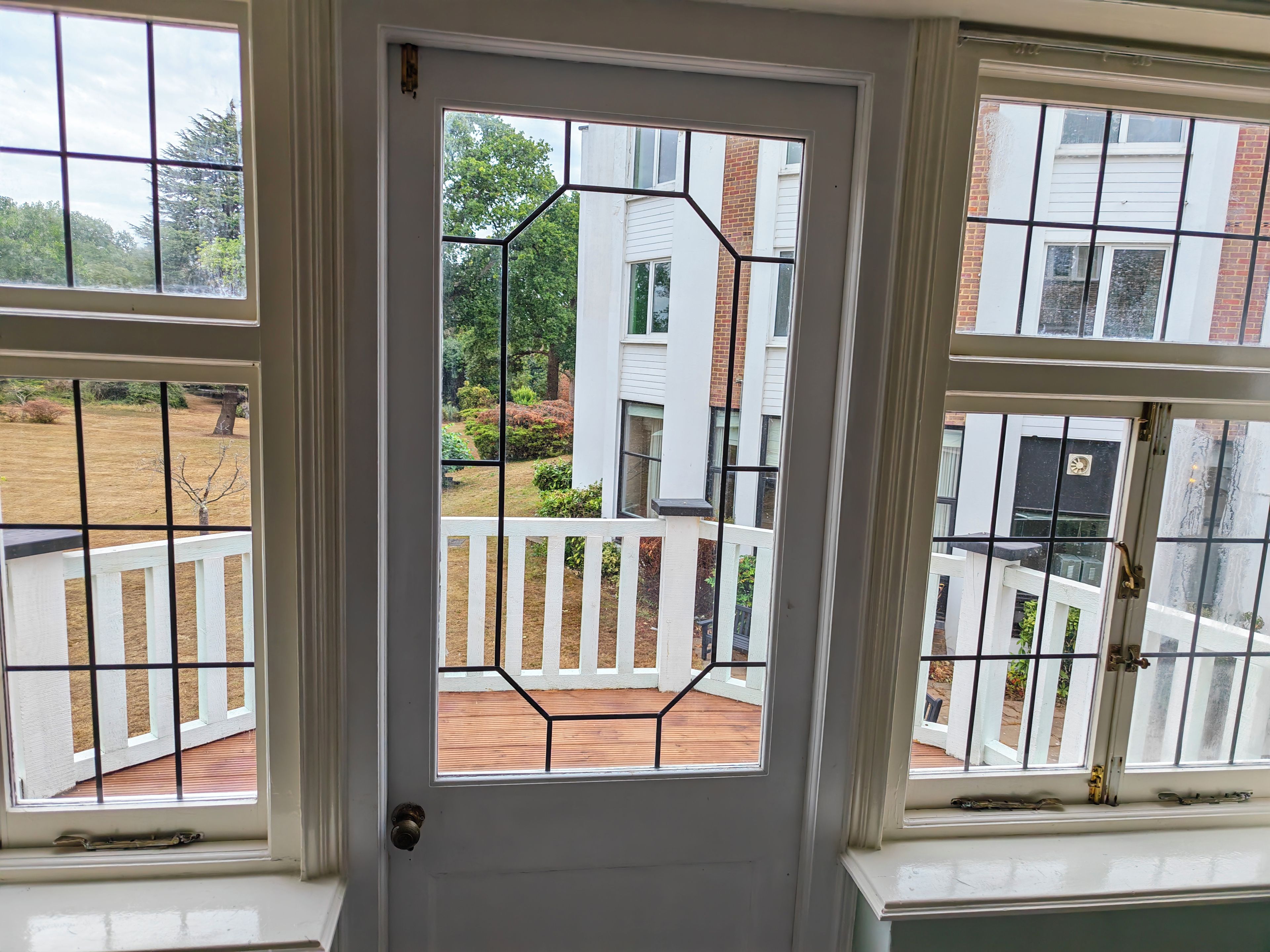 Puerta con diseño geométrico de vidrio, exterior blanco y vista a una terraza de madera y follaje.