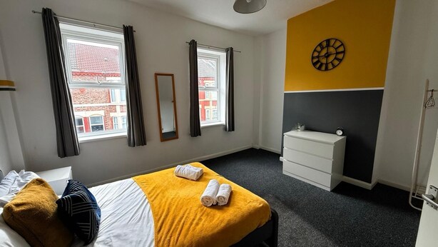 A hotel room with a bed, a bedside table, a dresser, and a wall clock. - House Sleeps 7 With Parking (Wallasey)