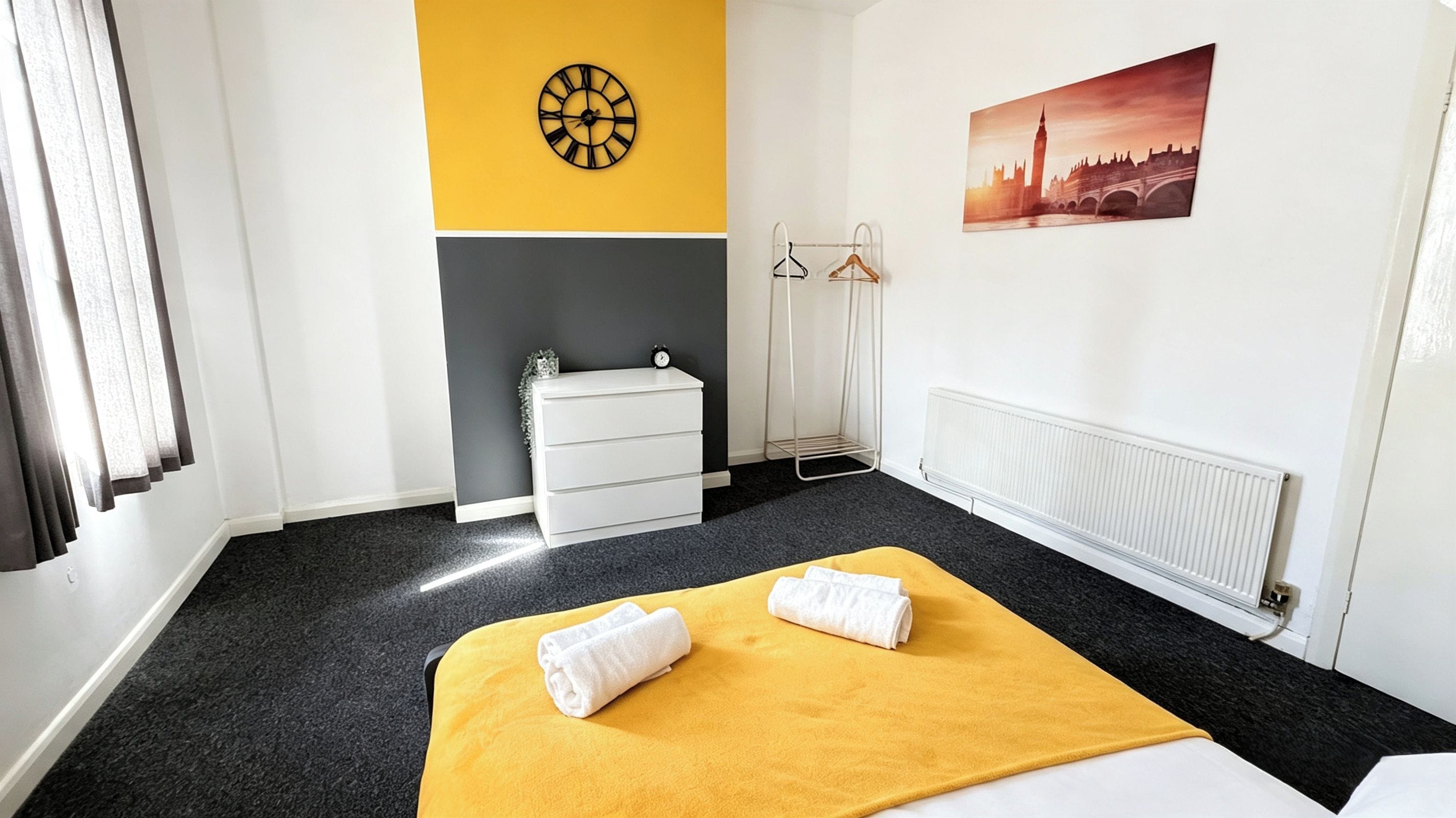 A hotel room with a bed, a dresser, a radiator, a clock, and a painting of a cityscape.