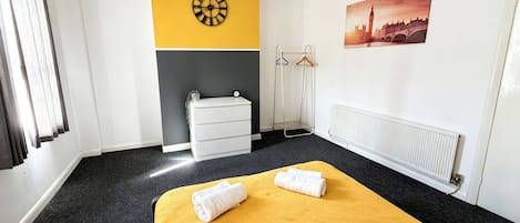 A hotel room with a bed, a dresser, a radiator, a clock, and a painting of a cityscape.