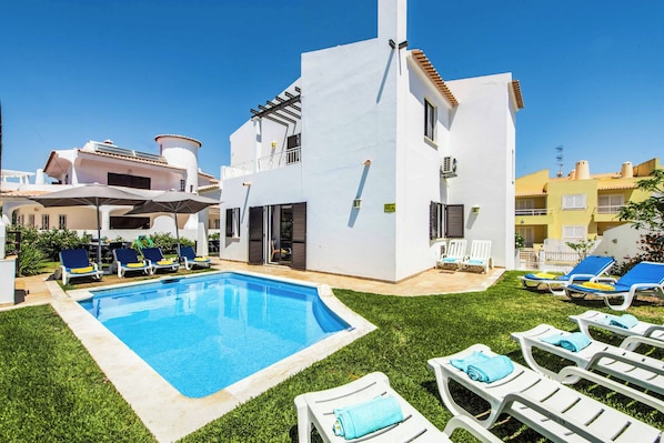 Zona de piscina amb una piscina blava i cristal·lina, gandules blanques i un edifici amb un balcó.