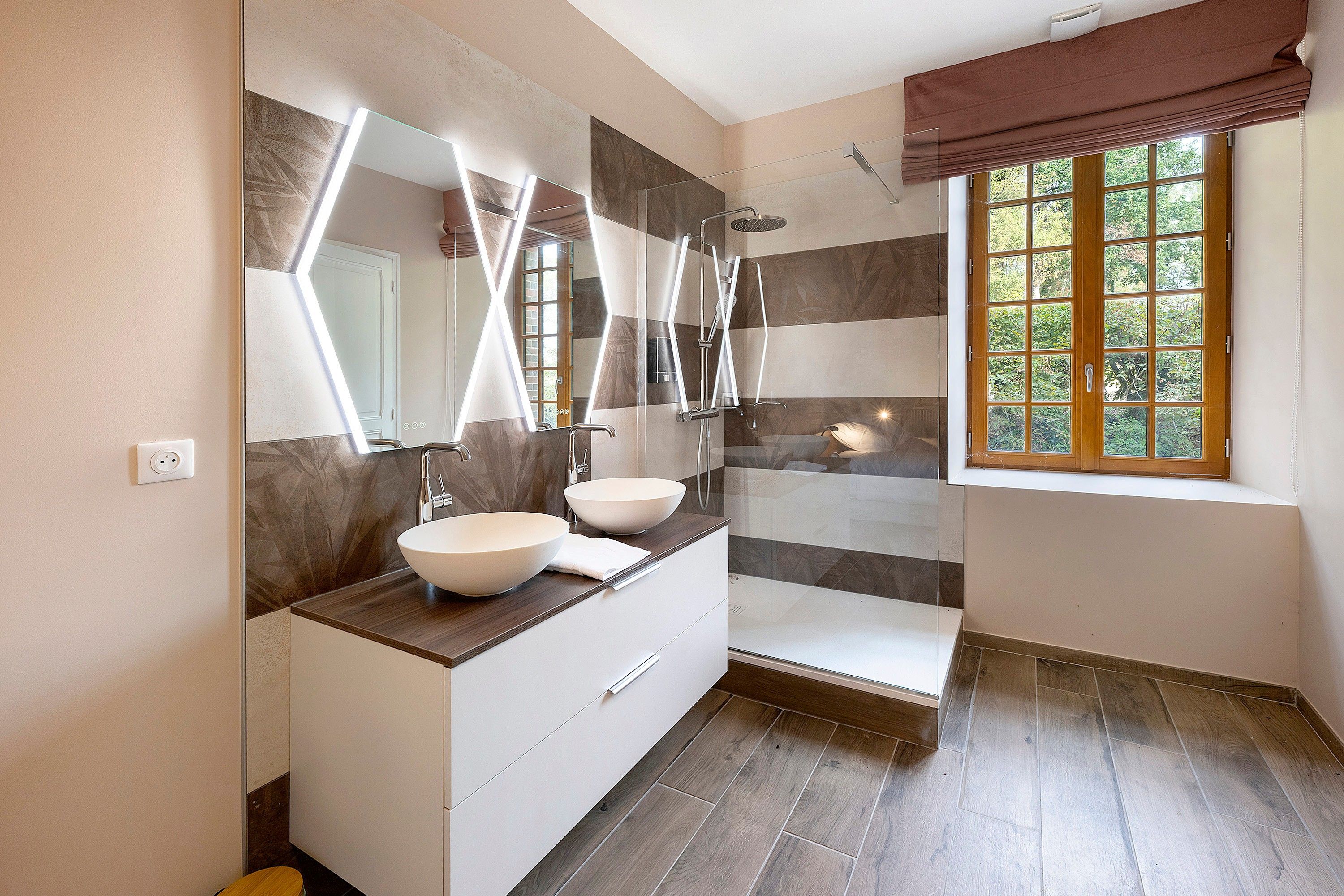 A modern bathroom with two sinks, a shower, and a window with a view of greenery.