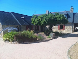 A stone building with a paved courtyard, a tree, and a clear blue sky. - Belle longère proche de la mer (Crozon)