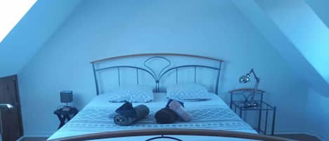 A single bed with a metal frame, two pillows, a bedside lamp, and a small table with a lamp.