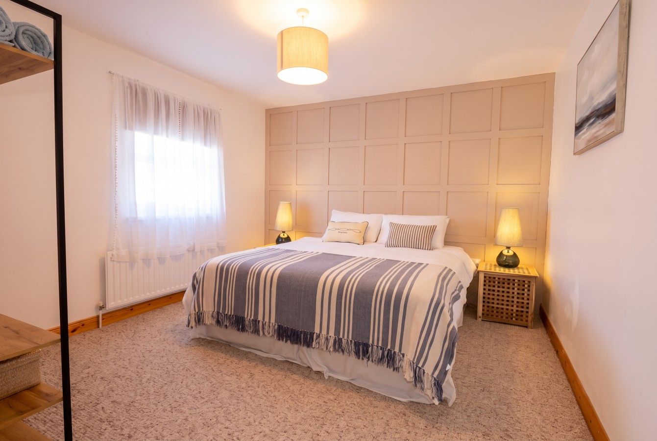 Quarto com cama, abajures, janela com cortinas, radiador e piso de madeira.