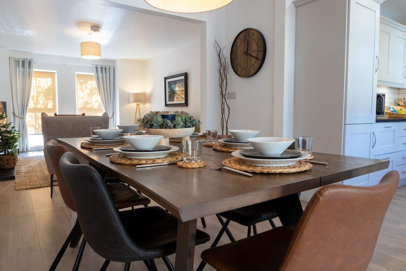 A dining area with a wooden table set for four, surrounded by chairs, with a clock on the wall and a plant on the table.