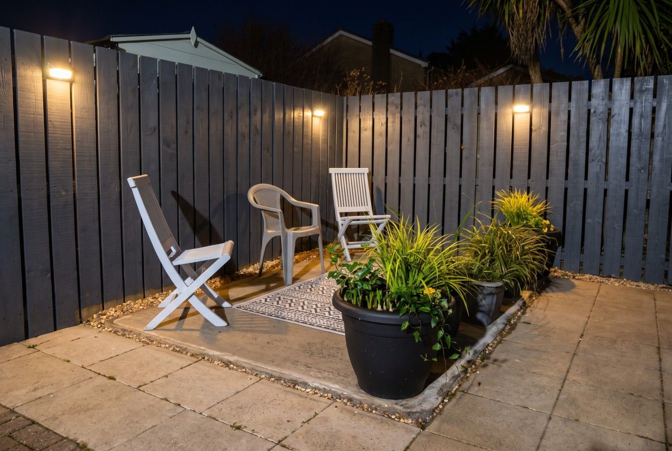A patio area with two white chairs, a black planter with plants, and a wooden fence.