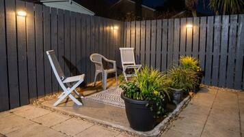 A patio area with two white chairs, a black planter with plants, and a wooden fence.