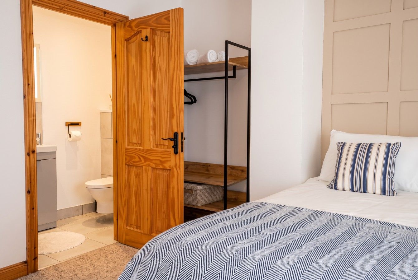 A bedroom with a bed, a wooden wardrobe, a bathroom with a toilet, and a shelf with rolled towels.