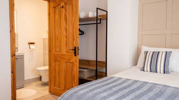 A bedroom with a bed, a wooden wardrobe, a bathroom with a toilet, and a shelf with rolled towels.