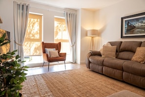 A living room with a brown sofa, a brown armchair, a floor lamp, and a large window with curtains. - 3-Bed Family House Sleeps 6 · Fireplace & Garden · Walk to Portstewart Golf Club (Co Londonberry)