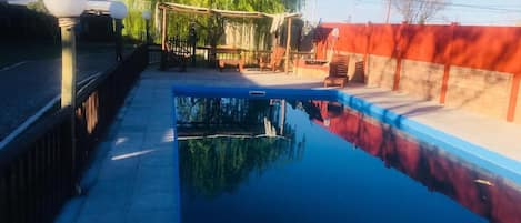A swimming pool with a wooden deck and lounge chairs, surrounded by a red wall and trees.