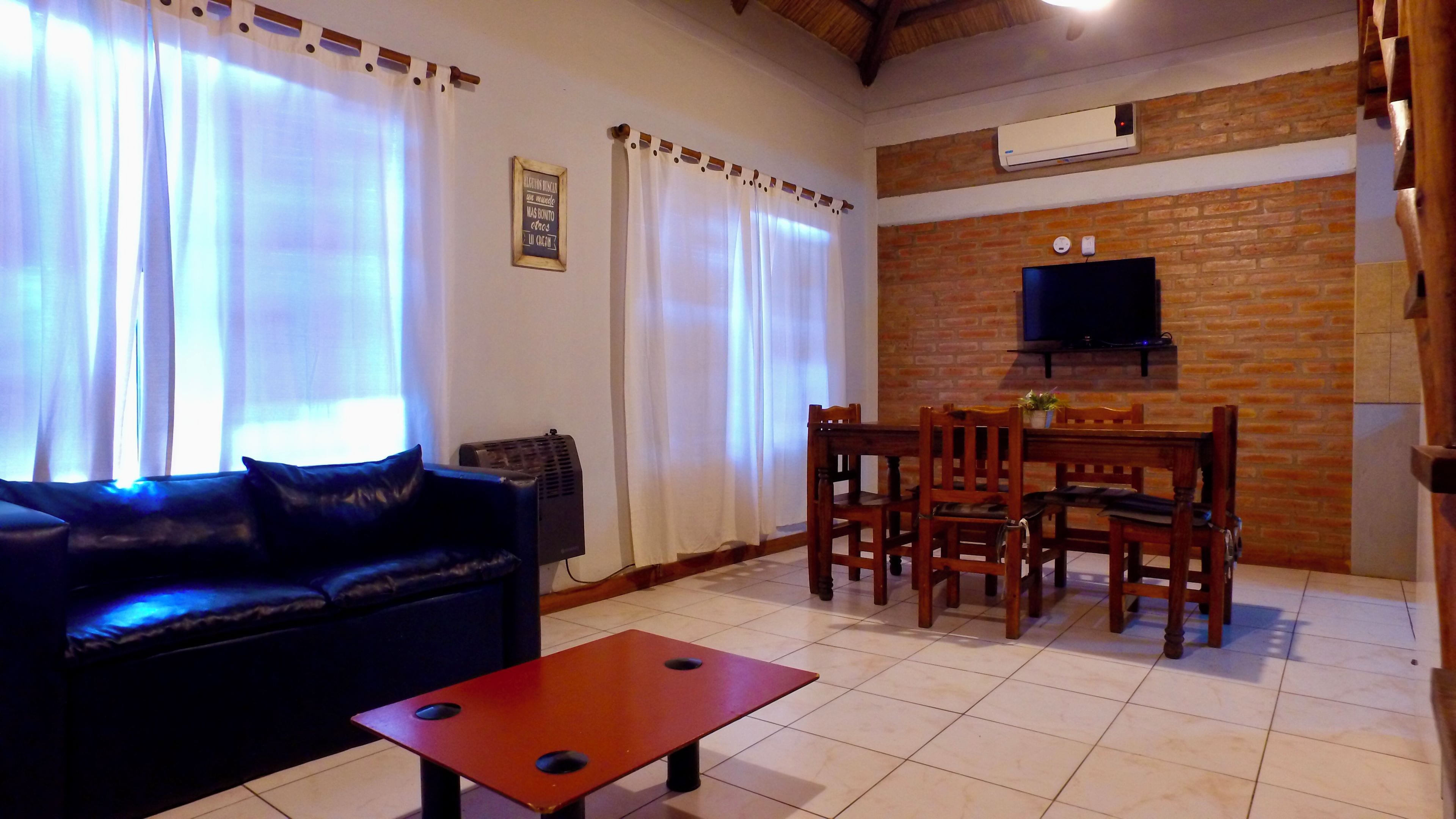 Uma sala de estar com sofá, mesa de centro, mesa de jantar com cadeiras, TV fixada em uma parede de tijolos e um aparelho de ar condicionado.