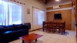 A living room with a sofa, a coffee table, a dining table with chairs, a TV mounted on a brick wall, and an air conditioning unit. - la aldea de san rafal (San Rafael)