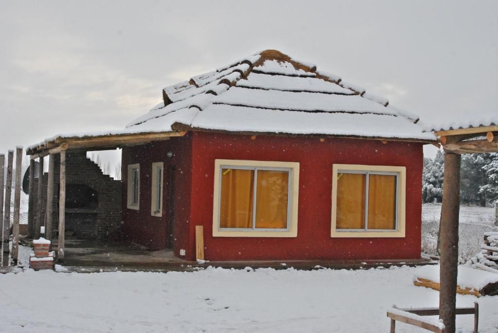 Un pequeño edificio rojo con techo nevado y dos ventanas con cortinas amarillas.