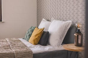 Ein ordentlich bezogenes Bett mit einem gemusterten Überwurf, einer Auswahl an Kissen und einem Nachttisch mit einer Vintage-Lampe.