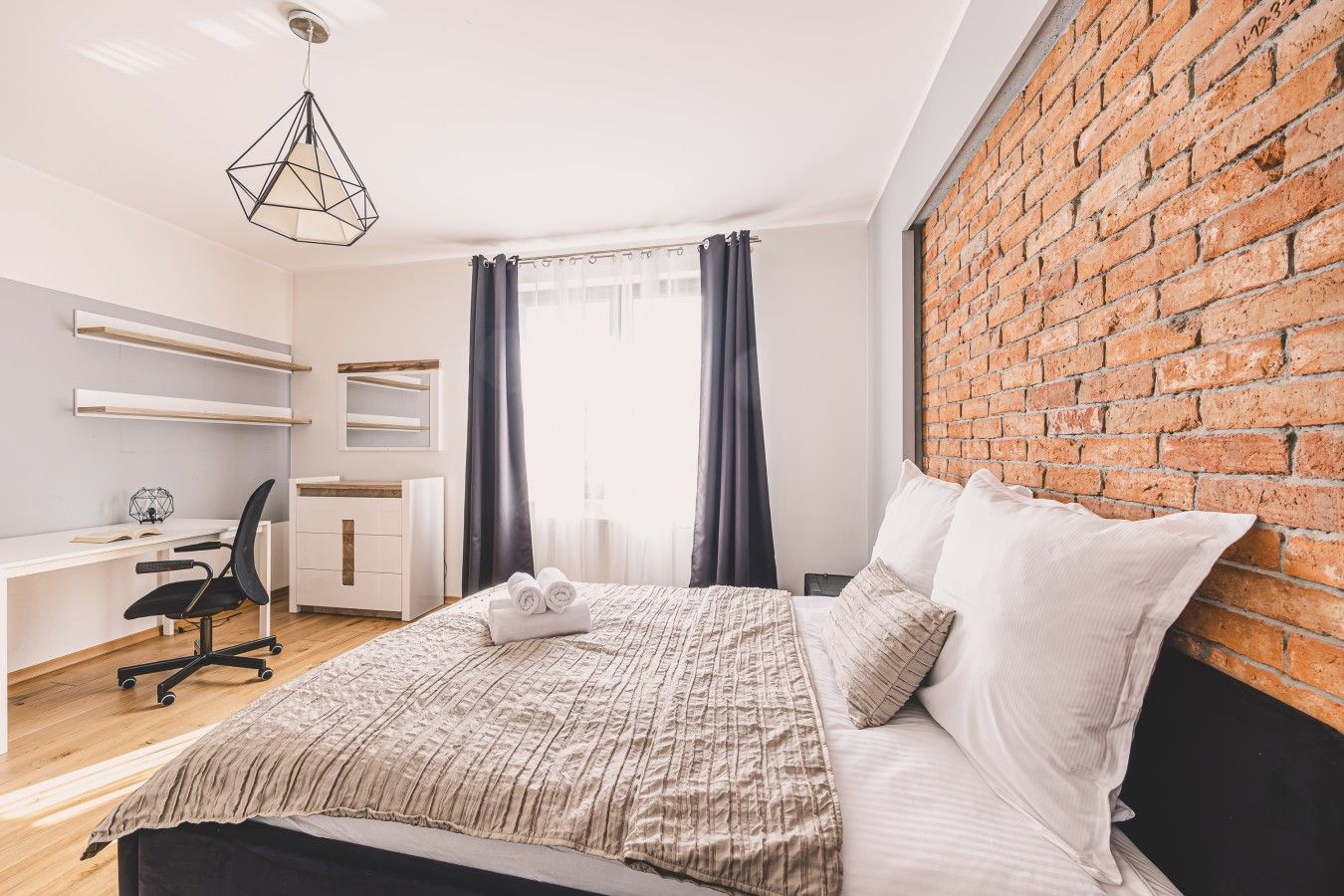 A modern bedroom with a brick wall, a bed with white bedding, a desk with a chair, and a hanging light fixture.