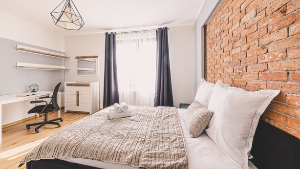 A modern bedroom with a brick wall, a bed with white bedding, a desk with a chair, and a hanging light fixture. - Dream 30 | Duży Loft z 2 Sypialniami | Bydgoszcz (Bydgoszcz)