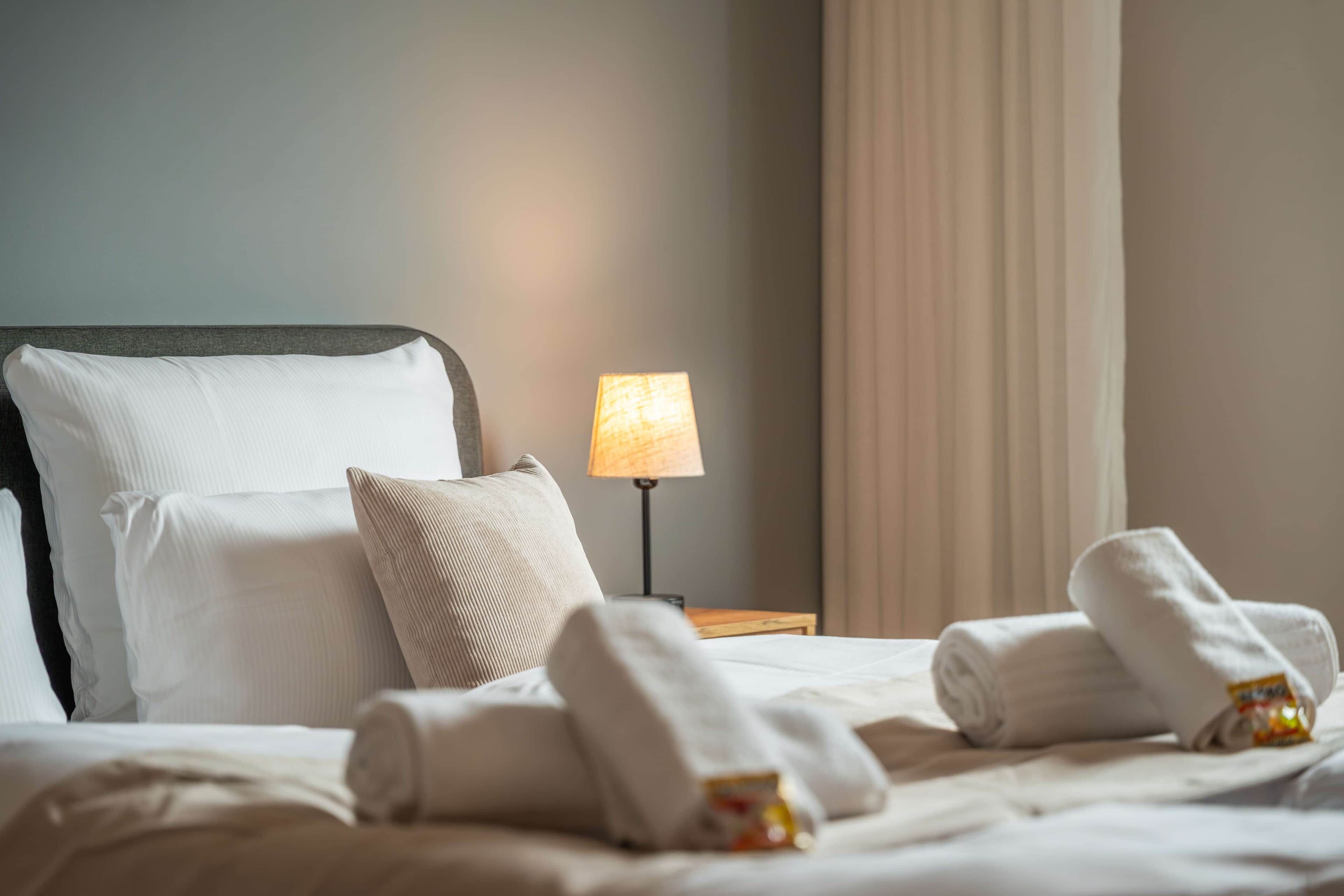 Ein ordentlich gemachtes Bett mit weißen Leinen und aufgerollten Handtüchern, eine Nachttischlampe und ein Vorhang.