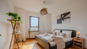 Ein modernes Schlafzimmer mit einem Bett, Nachttischen, einer Hängeleuchte, einem Fenster mit Blick und einer Pflanze auf einem Holzständer.