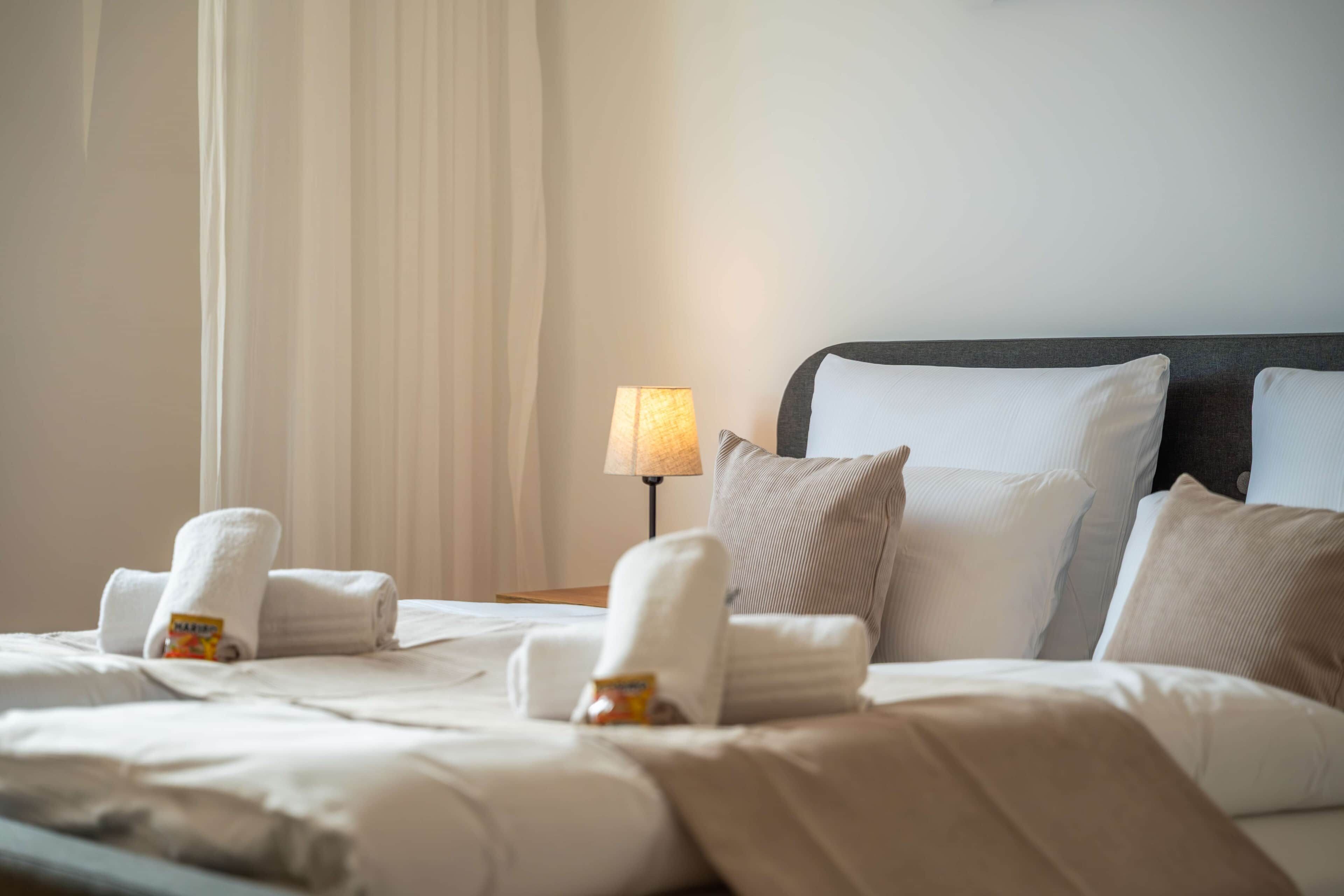 Una cama bien hecha con almohadas y una toalla enrollada, una lámpara de noche y una ventana con cortinas.