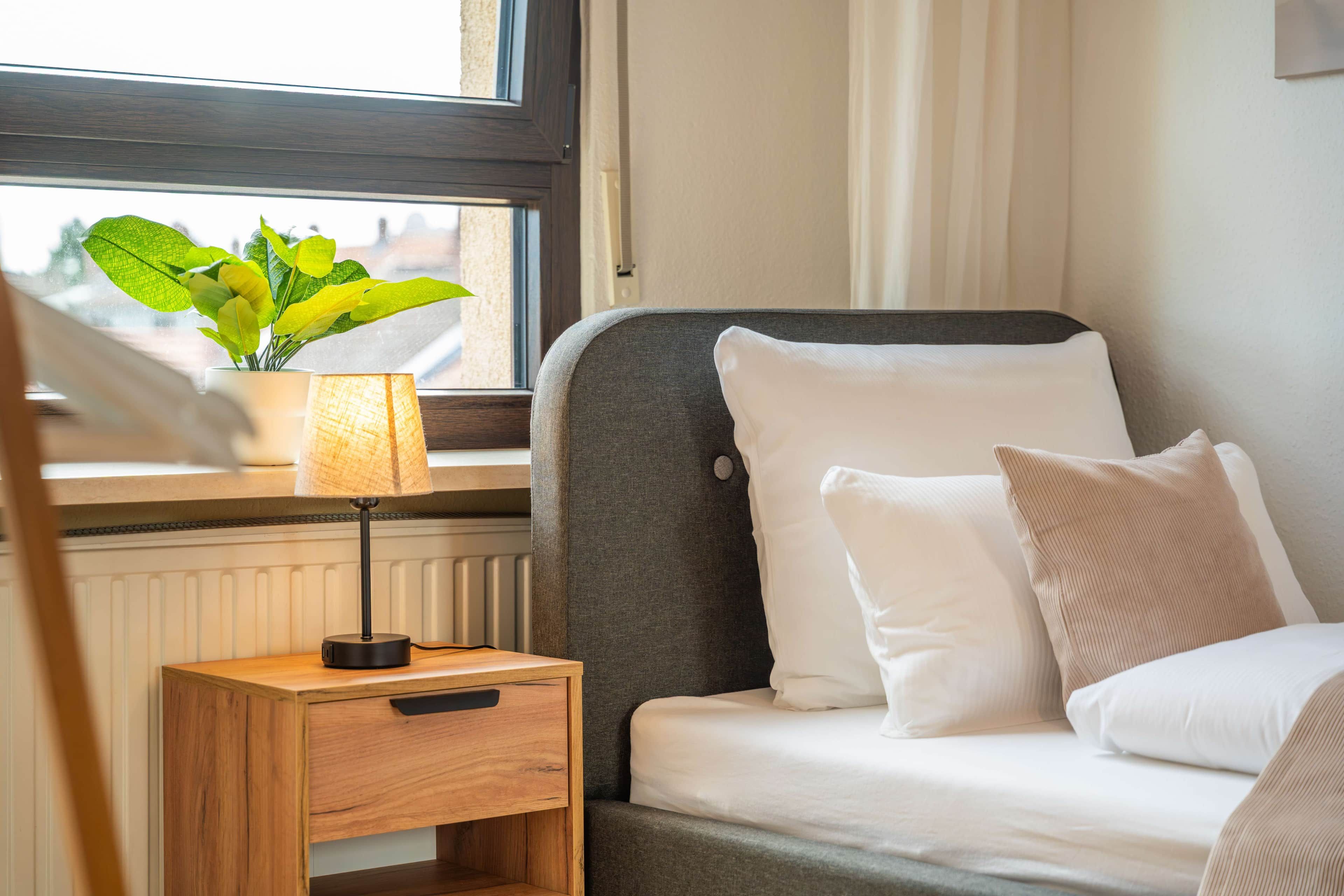 A neatly made bed with white bedding, a small bedside table with a lamp, and a window with a view of greenery outside.