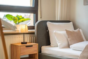 A neatly made bed with white bedding, a small bedside table with a lamp, and a window with a view of greenery outside.