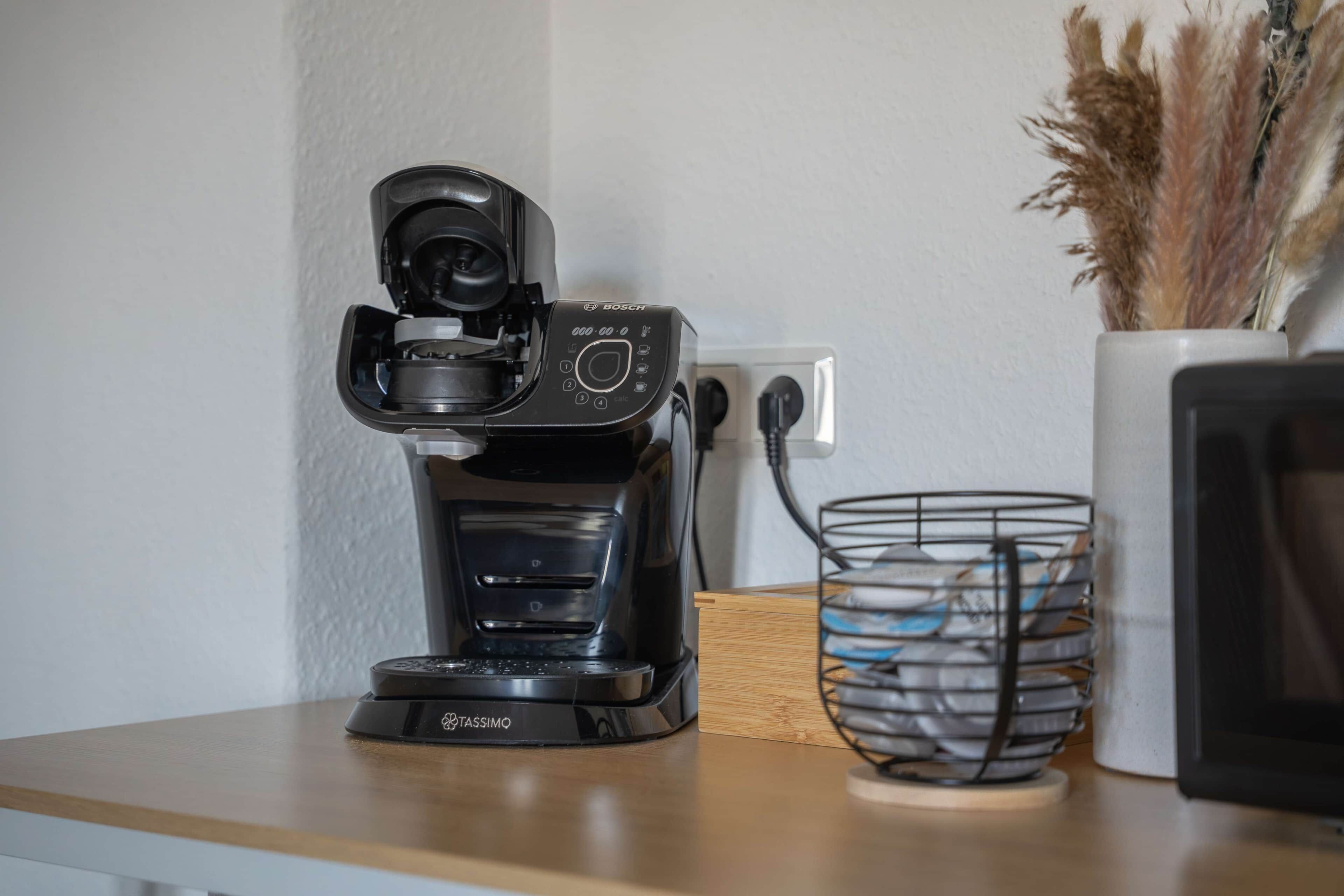Eine Kaffeemaschine auf einer Holztischplatte, ein Steckdosenanschluss und im Hintergrund ein Korb mit Gegenständen.