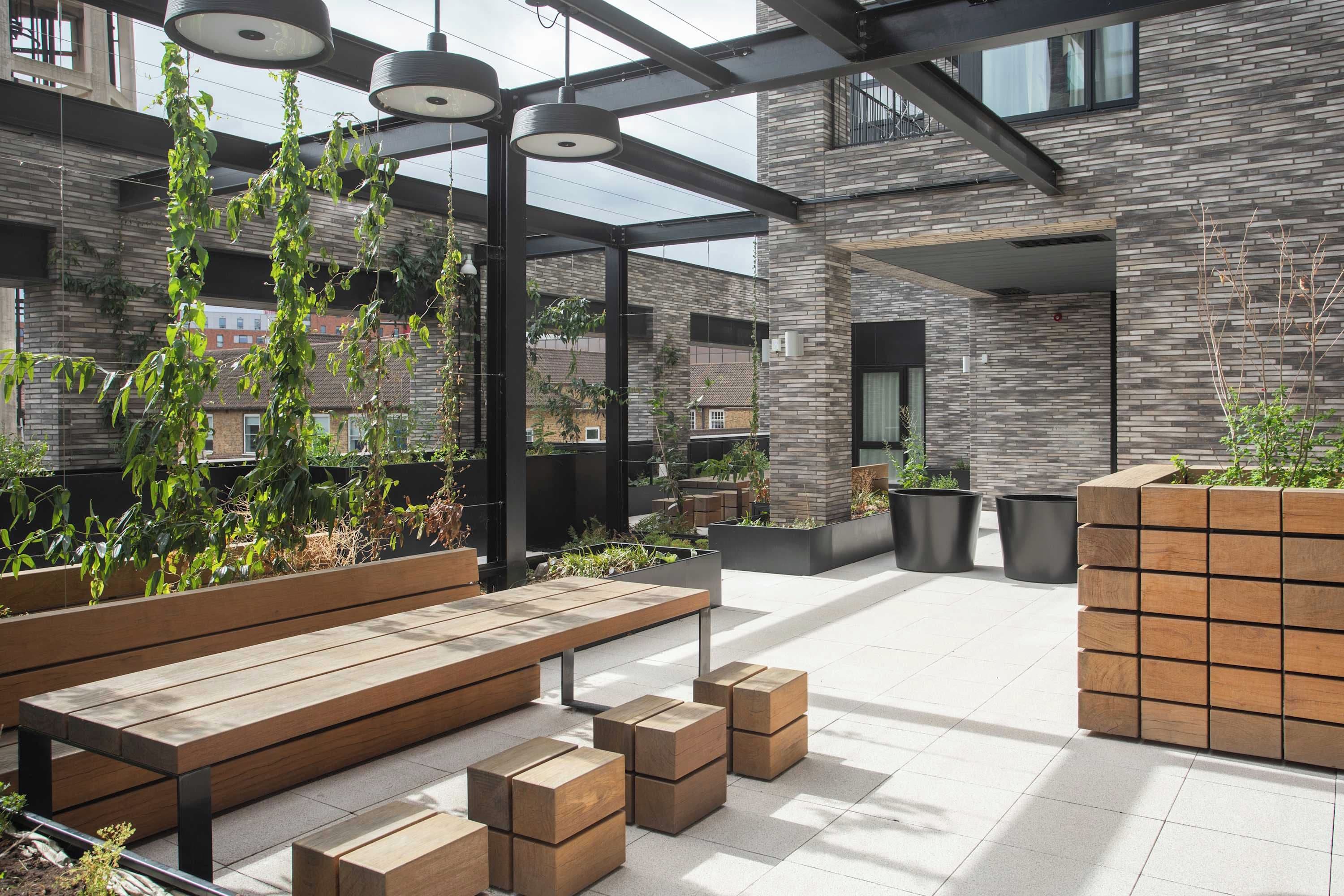 An outdoor seating area with wooden benches and planters, a modern building with a stone facade, and hanging pendant lights.