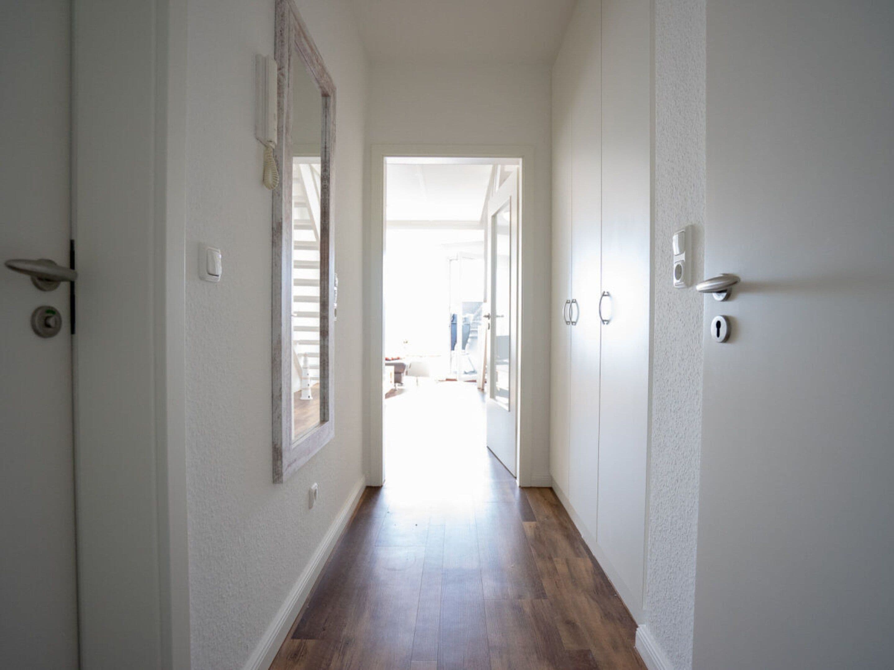 A hallway with a wooden floor, white walls, and a door leading to another room.