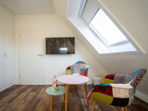 Ein Zimmer mit schrägen Wänden, einem an der Wand befestigten Fernseher, einem runden Tisch mit zwei Stühlen und einem kleinen Dekoartikel auf dem Boden.