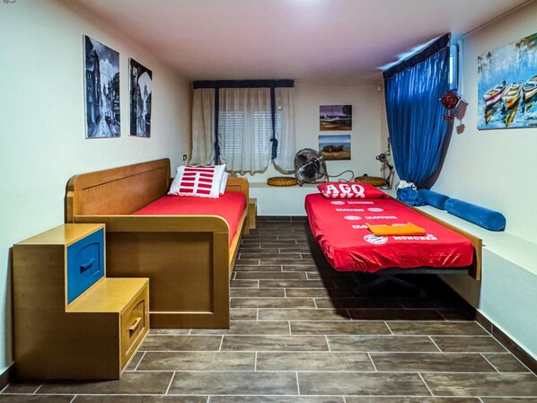 A room with two beds, a small desk, a fan, and a window with curtains.