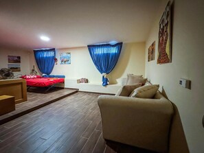 Una habitación con sofá, cama con ropa de cama roja, una mesita y una ventana con cortinas azules.
