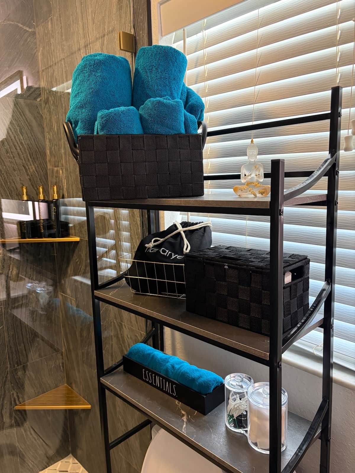 A black metal shelf unit with a blue towel basket, a black mesh bag, and a glass jar on the lower shelf.