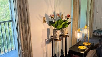 A room with a bed, a bedside table with a lamp, a window with curtains, and a view of greenery outside.
