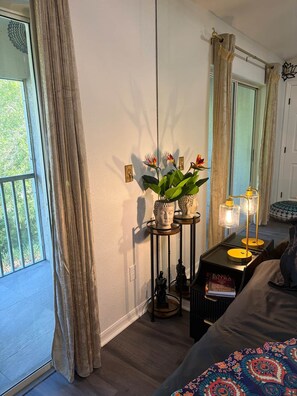 Ein Zimmer mit einem Bett, einem Nachttisch mit Lampe, einem Fenster mit Vorhängen und Blick ins Grüne.