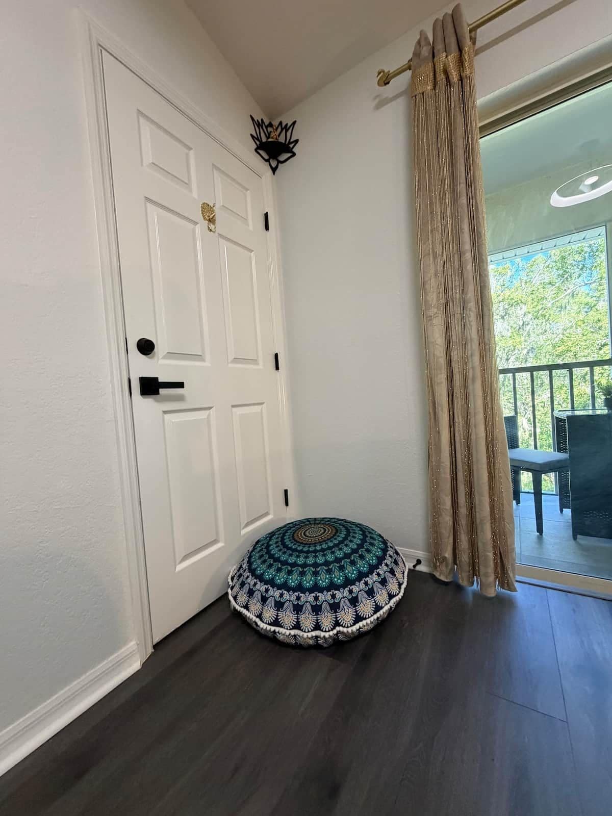 A room with a white door, a round patterned cushion, and a view of an outdoor area through a glass door.