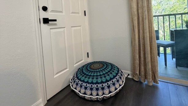 A room with a white door, a round patterned cushion, and a view of an outdoor area through a glass door. - Paradise Goddess at Paradise Lakes Nudist Resort (Land O' Lakes)